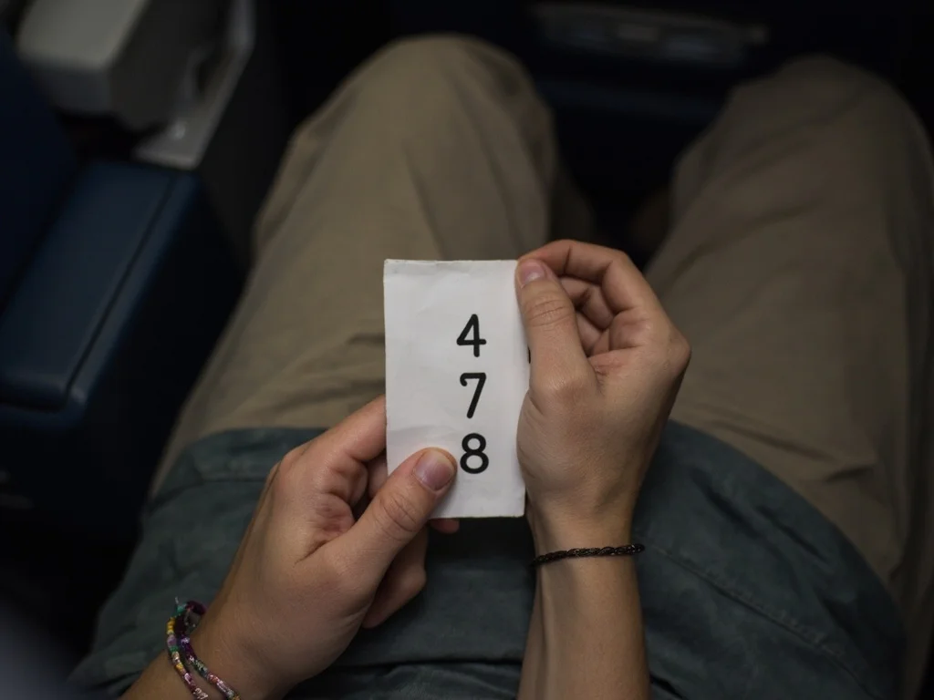 Mãos no colo de um passageiro: uma segura um papel com os números 4, 7 e 8, a outra usa uma pulseira trançada.